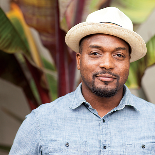 a black man looks at the camera softly smiling wearing a light blue button down collared shirt and a light cream colored brimmed hat against a tropical plant background