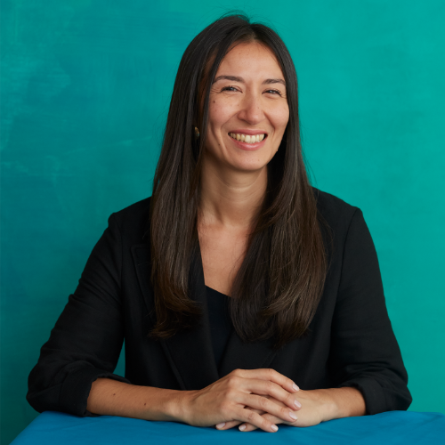 a professional headshot of an asian woman with long dark hair smiling at the camera, she is sitting against a blue/green background wearing a black shirt with 3/4 length sleeves and her hands are folded across eachother